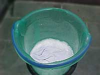 Foto de uma pessoa despejando, em uma tampinha, um pouco de álcool em um balde com água e sabão. O objetivo é clarear a roupa branca