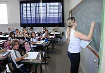 Foto de 
uma professora escrevendo no quadro negro e os alunos copiando matéria de suas carteiras