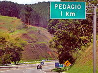 Foto da estrada com um carro e uma placa de ped?gio escrita que h? ped?gio a 1Km